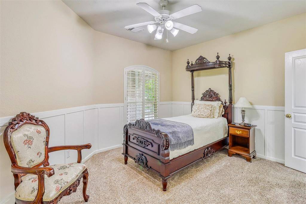 This second generously sized secondary bedroom combines comfort and charm, featuring a high ceiling with a ceiling fan, soft plush carpeting, and elegant wainscoting accents that add a touch of classic detail. A large window fitted with plantation shutters allows natural light to pour in while offering privacy and style.