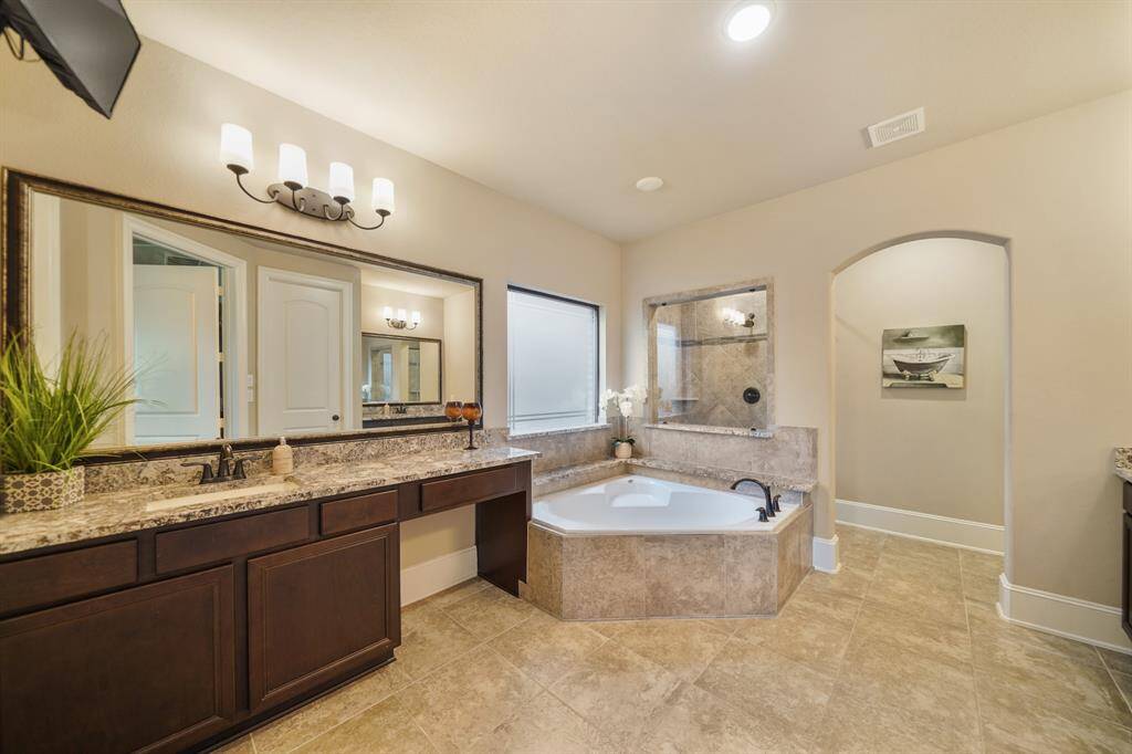 This elegant primary bathroom features a large soaking tub, dual sinks with granite countertops, and ample cabinetry