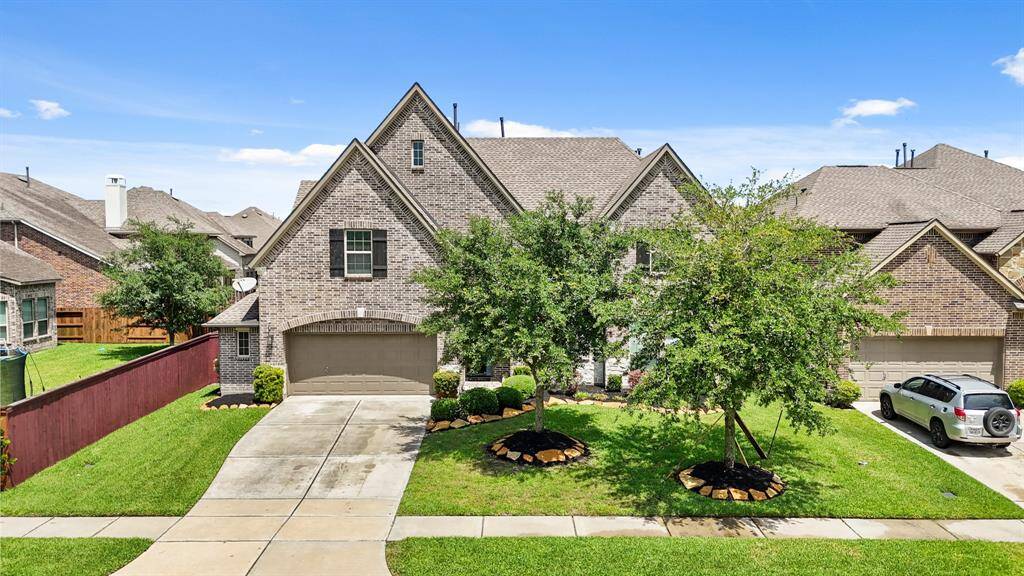 This is a charming two-story brick home with gabled roofs and a double garage. The front yard features a well-maintained lawn and mature trees, offering a welcoming curb appeal in a suburban neighborhood.