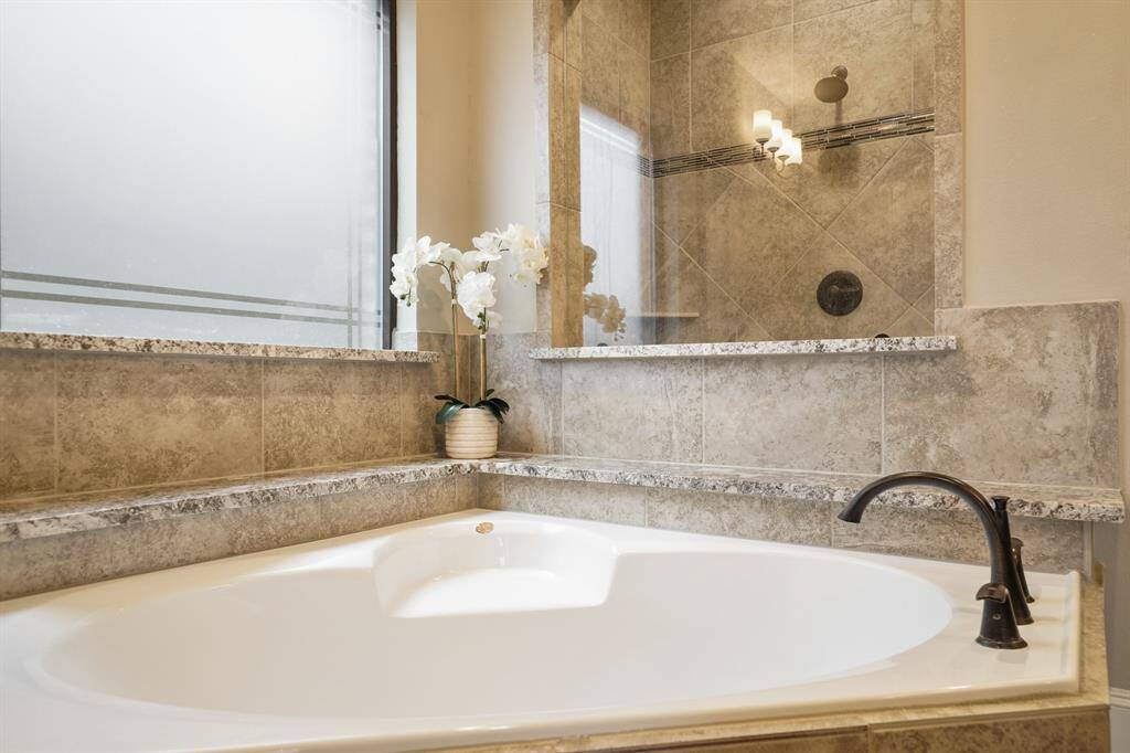 This bathroom features a luxurious corner bathtub with elegant tile surround, a granite ledge, and a modern faucet. A large window provides natural light, and a stylish shower is visible in the background.