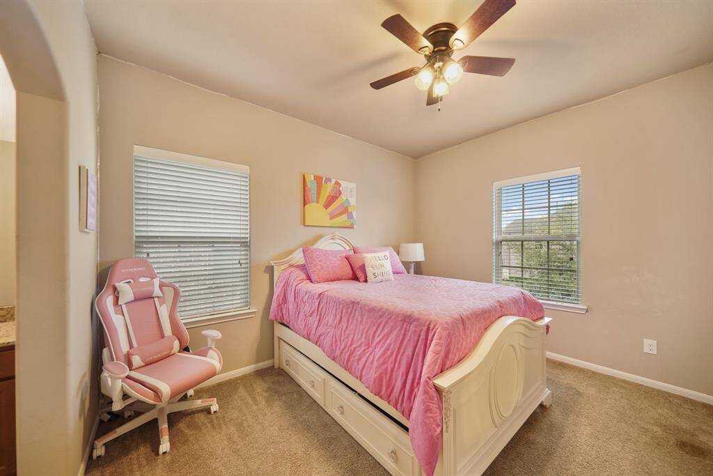 Cozy bedroom with soft beige walls and carpet, featuring a ceiling fan, two windows for natural light. Ideal for a comfortable and bright personal space.