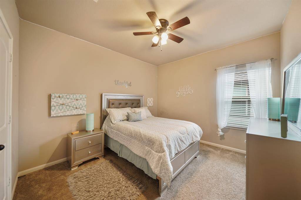 This cozy bedroom features neutral tones with a plush carpet, a ceiling fan, and abundant natural light creating a comfortable and inviting space.