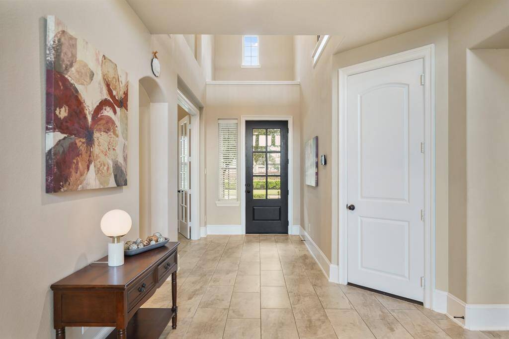 A bright, welcoming foyer with light wood flooring, neutral walls, and a black front door. Natural light streams through upper windows, enhancing the open feel.