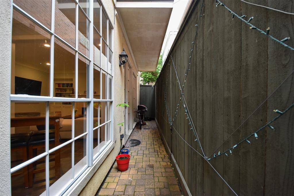 Well-lit side path of the home, featuring large windows providing a view into the family room, a secure high fence on one side, and a paved walkway offering a private access point