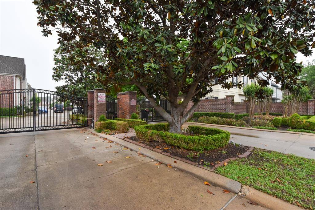 Gated community entrance featuring a wrought-iron gate, guest parking, well-maintained landscaping with mature trees in an established neighborhood