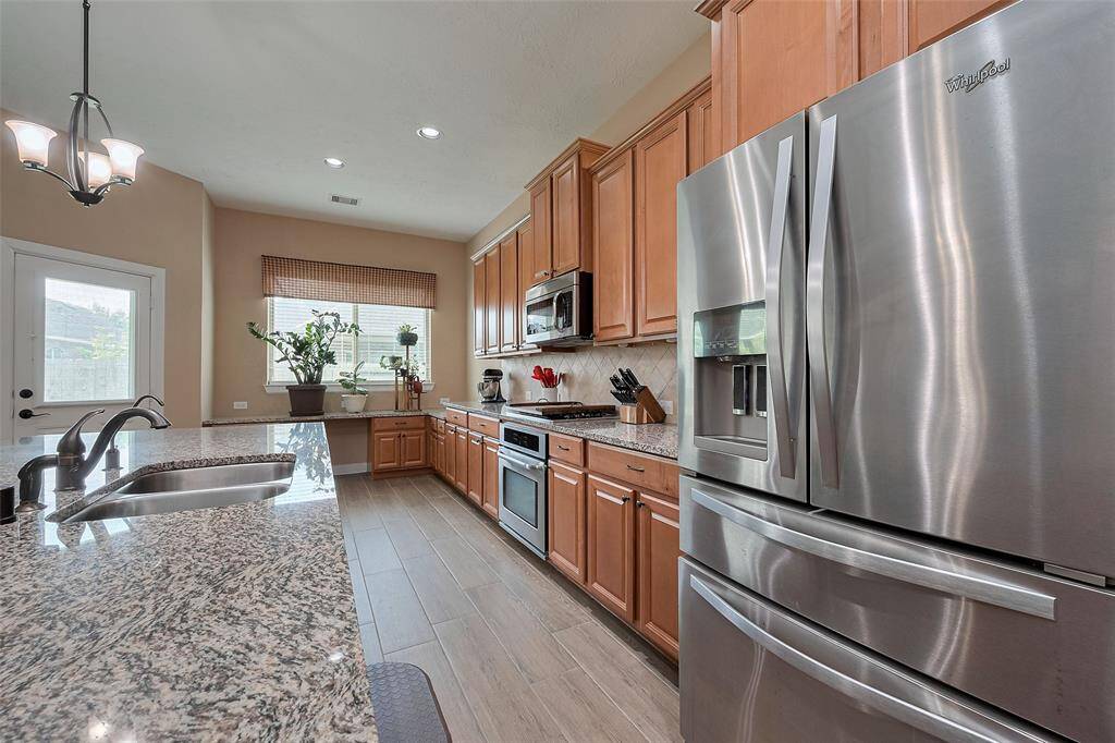 This kitchen features modern stainless steel appliances, granite countertops, and ample wooden cabinetry. It has a double sink, pendant lighting, and a bright, inviting atmosphere with large windows and a door leading outside.