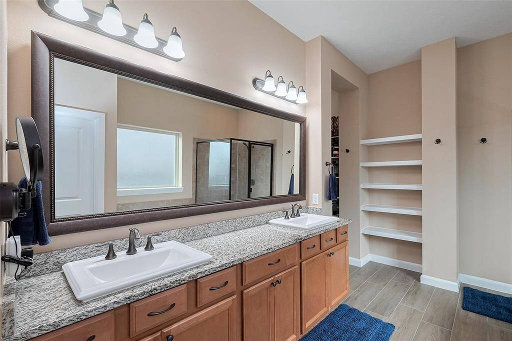 The primary ensuite bathroom features a spacious double vanity with curtured marble countertops, ample storage, a large mirror, and modern lighting. It includes a glass-enclosed shower and built-in shelving for convenience.
