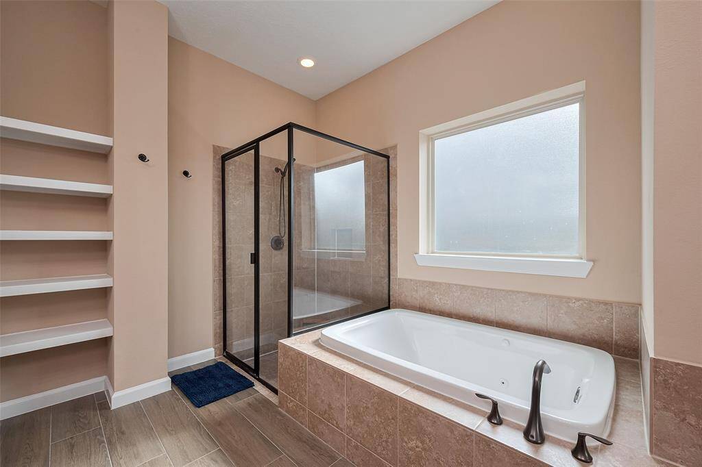 The primary bathroom features a modern design with a spacious glass-enclosed shower, a luxurious built-in bathtub, and sleek tile flooring. It includes open shelving for storage and a large frosted window for natural light and privacy.
