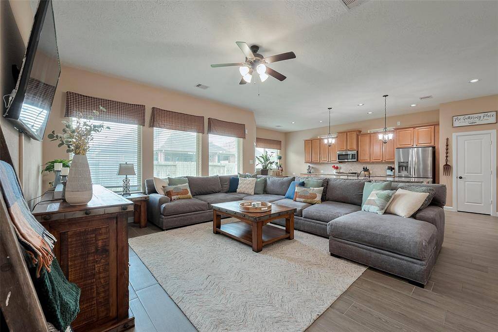 The room is bright with large windows, a modern ceiling fan. The kitchen boasts stainless steel appliances, wooden cabinetry, and stylish pendant lighting, seamlessly connecting to the living area for easy entertaining.