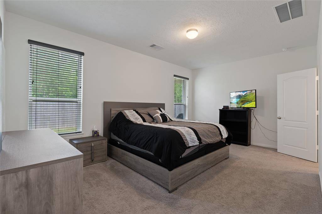 This bedroom is spacious and bright, featuring neutral carpeting, two windows with blinds, and modern furniture. It includes a large bed, a dresser, and a TV on a stand, creating a comfortable and inviting atmosphere.