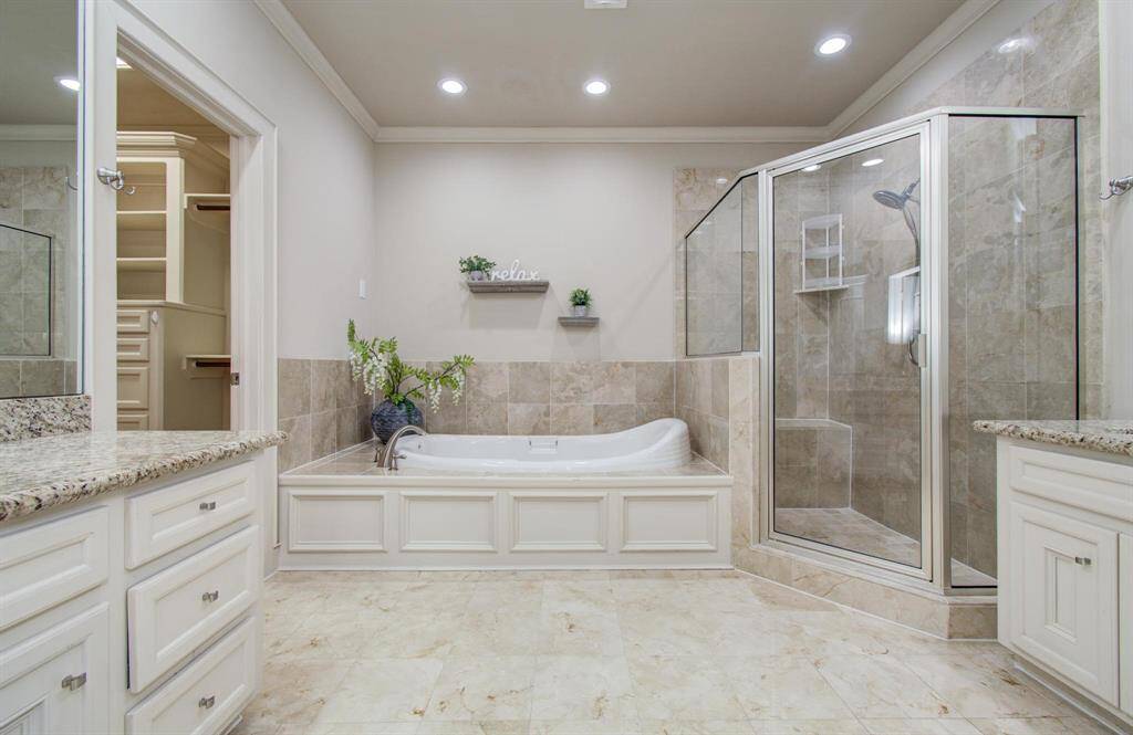 The primary bathroom features a large granite countertop with double sinks, a glass-enclosed shower, and a luxurious soaking tub with decorative shelving. The neutral tones and recessed lighting create a relaxing, elegant atmosphere.