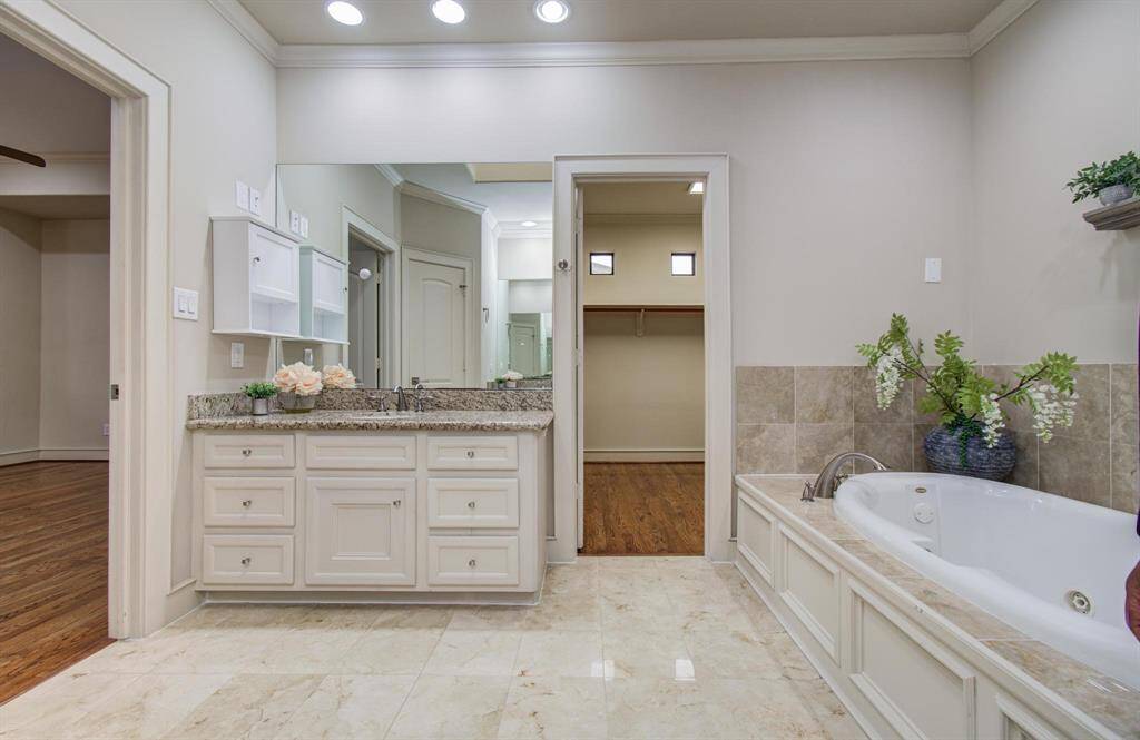 The primary bathroom features a large granite countertop with double sinks, a glass-enclosed shower, and a luxurious soaking tub with decorative shelving. The neutral tones and recessed lighting create a relaxing, elegant atmosphere.