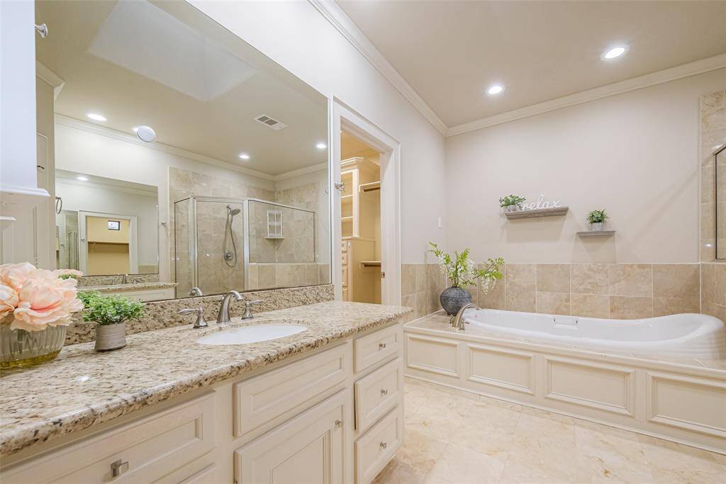 The primary bathroom features a large granite countertop with double sinks, a glass-enclosed shower, and a luxurious soaking tub with decorative shelving. The neutral tones and recessed lighting create a relaxing, elegant atmosphere.