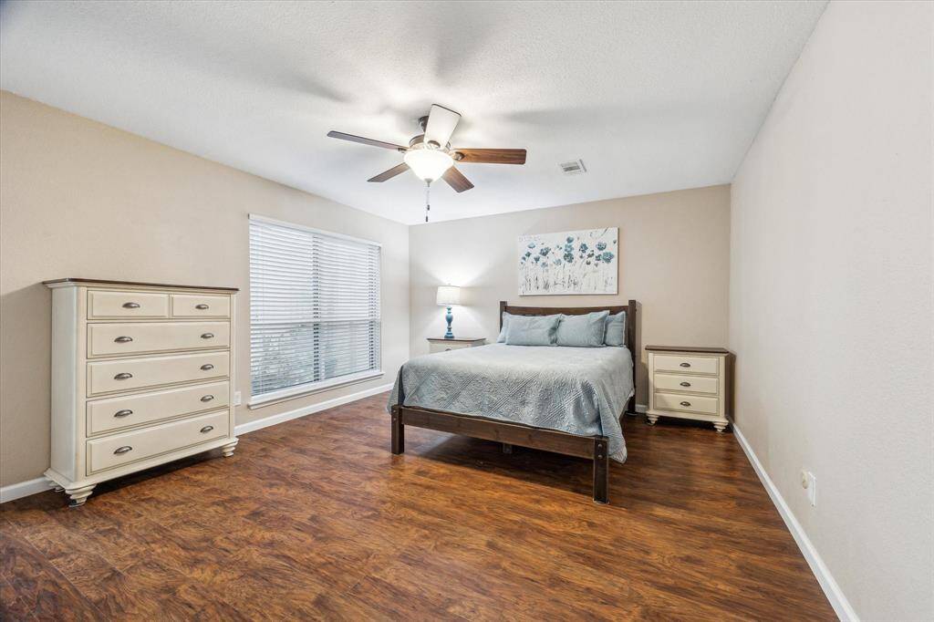The primary bedroom has warm wood flooring, neutral walls, and natural light from a large window.