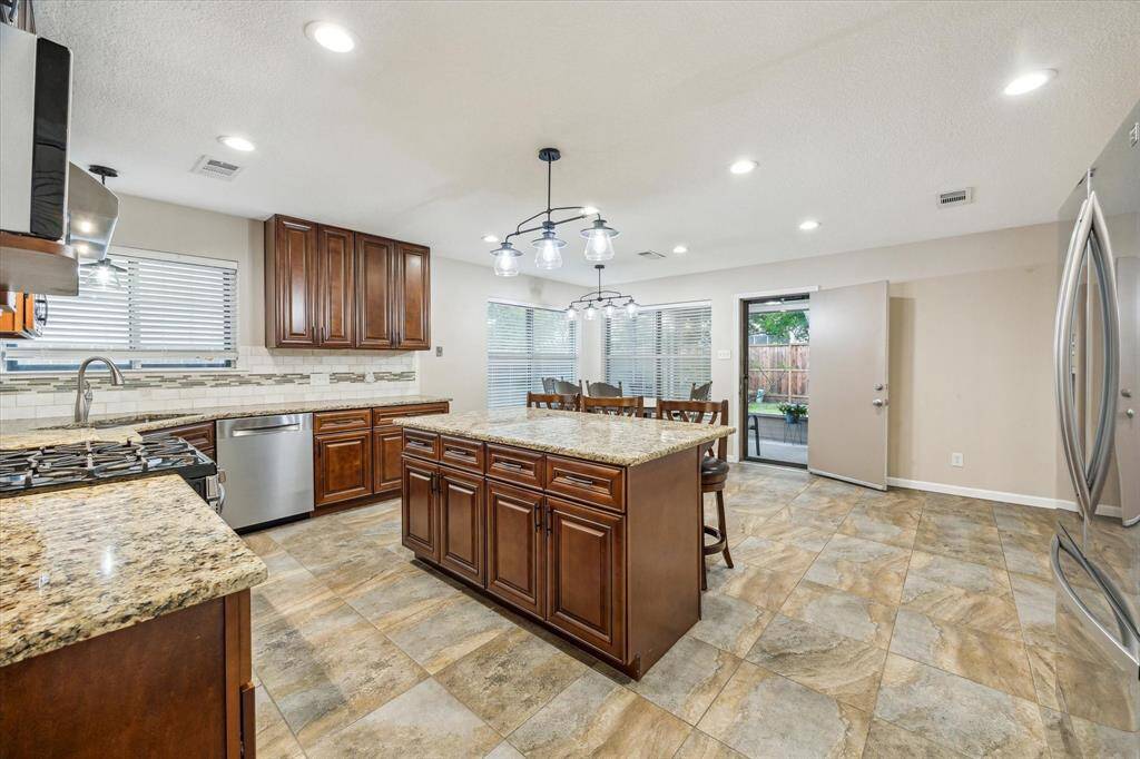 Spacious kitchen with modern amenities featuring granite countertops, stainless steel appliances, and rich wooden cabinetry. The open layout includes a central island with seating, tiled flooring, and a patio door leading out to the sunroom.