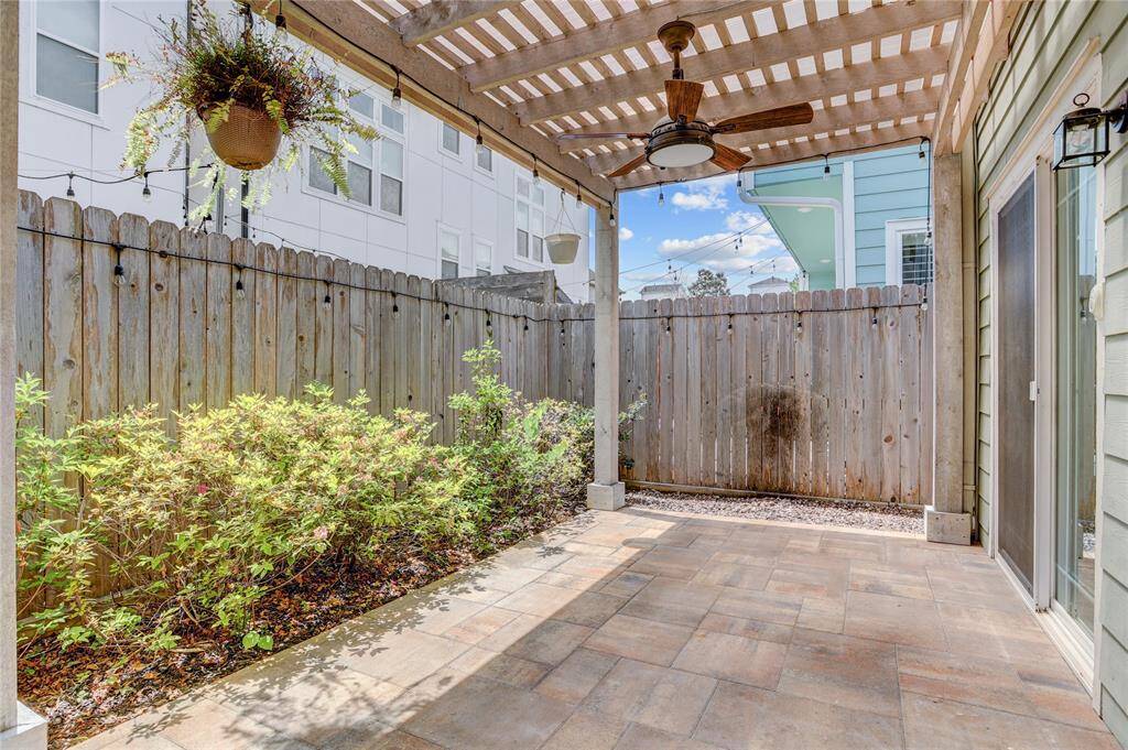 Another view of the backyard shows the covered patio, privacy fence, and landscaped border. The space includes a ceiling fan, hanging lights, and direct access to the home through sliding doors.