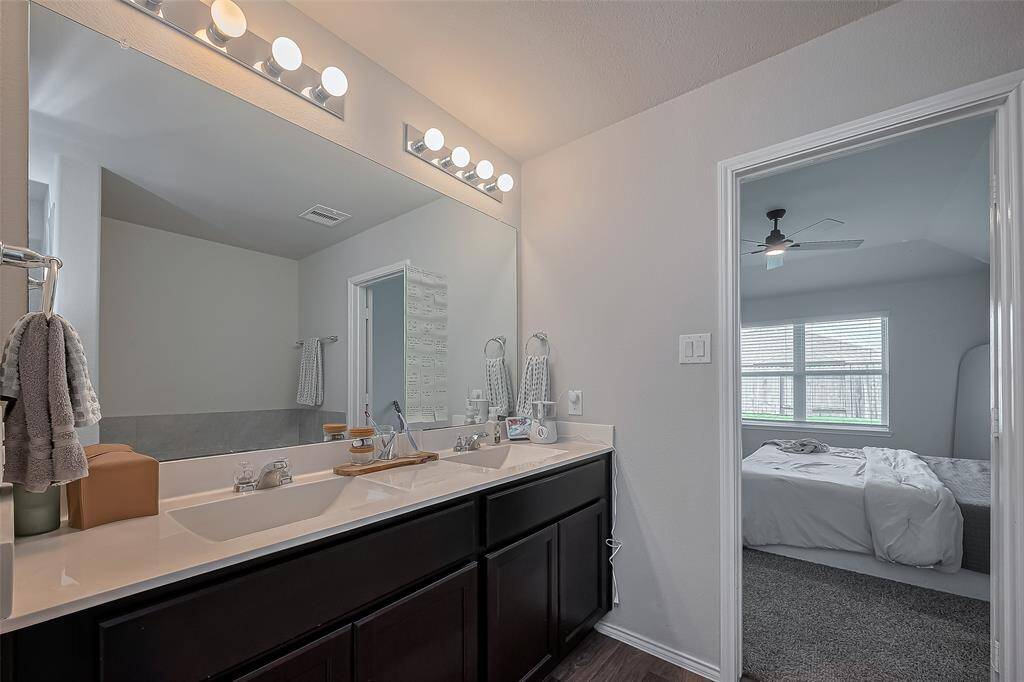 Primary suite includes a double vanity and ample counter space, enhanced by bright vanity lighting. It opens into a cozy bedroom with large windows and a ceiling fan, creating an inviting atmosphere.