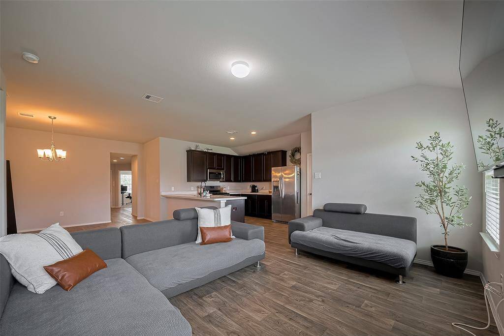 This photo showcases a spacious open-concept living area with modern finishes. The room features sleek wood flooring, a cozy seating arrangement, and ample natural light from large windows. The kitchen is visible with expresso cabinetry and stainless steel appliances, creating a seamless flow between spaces. Ideal for comfortable living and entertaining.