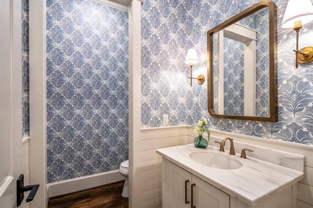 The powder room blends charm and sophistication with marble countertops, brushed gold fixtures, and a floral wallpaper that feels both timeless and on trend. The sink area seen here offers future elevator capability. It can easily be converted to an elevator space, syncing up with the second floor shaft. The sink would then go on the other side of the lavatory (currently a wet bar).