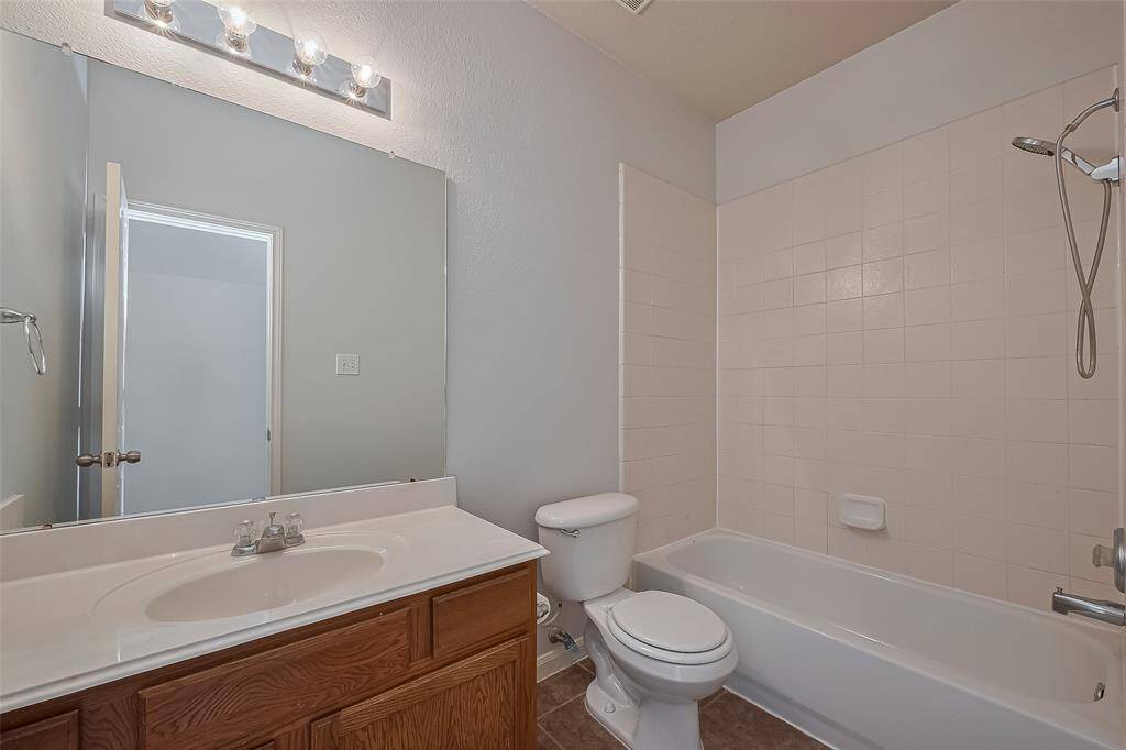 This photo shows a clean, simple bathroom featuring a bathtub with a shower, a toilet, and a single-sink vanity with a large mirror and overhead lighting. The walls are a neutral color, complementing the light tile and wood cabinetry.