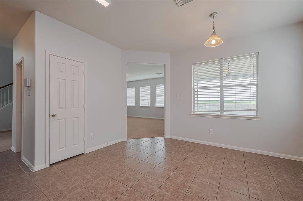This photo shows a bright dining area with tiled flooring, a single hanging light fixture, and large windows offering plenty of natural light. There's a doorway leading to a carpeted living space and a door to the side, possibly for storage or a pantry.