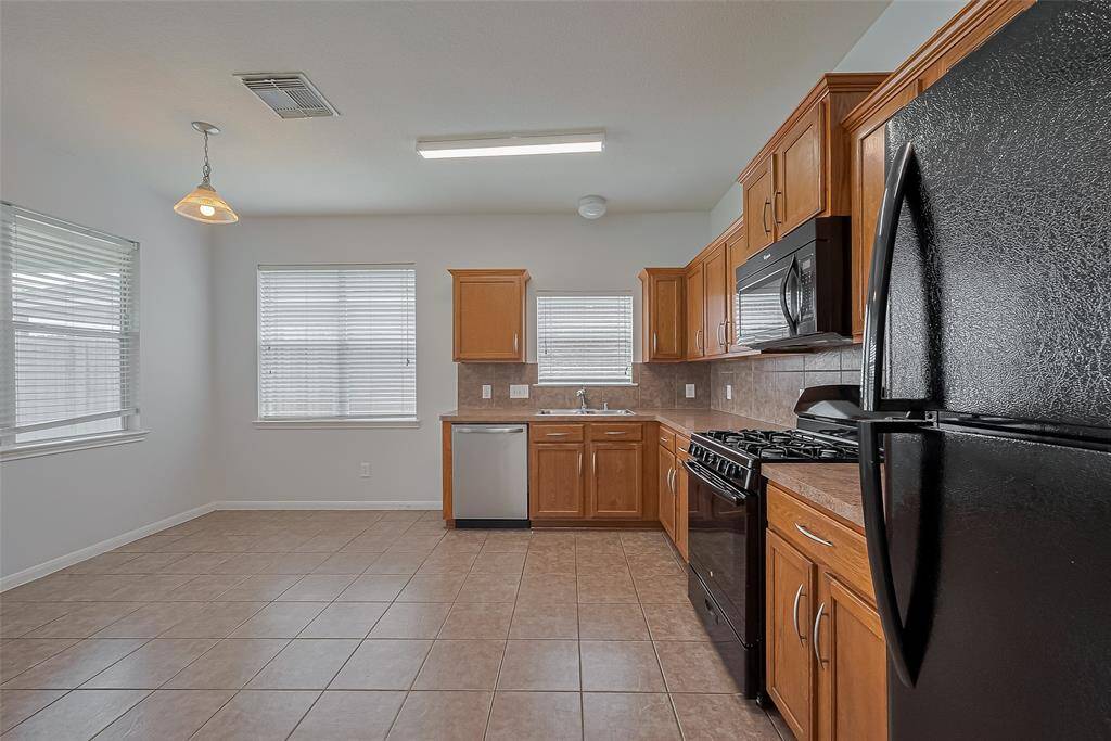 This spacious kitchen features ample natural light, tiled flooring, and wooden cabinetry. It includes modern appliances with a black finish, a large refrigerator, and a gas stove. The open layout offers a cozy dining area under a pendant light.