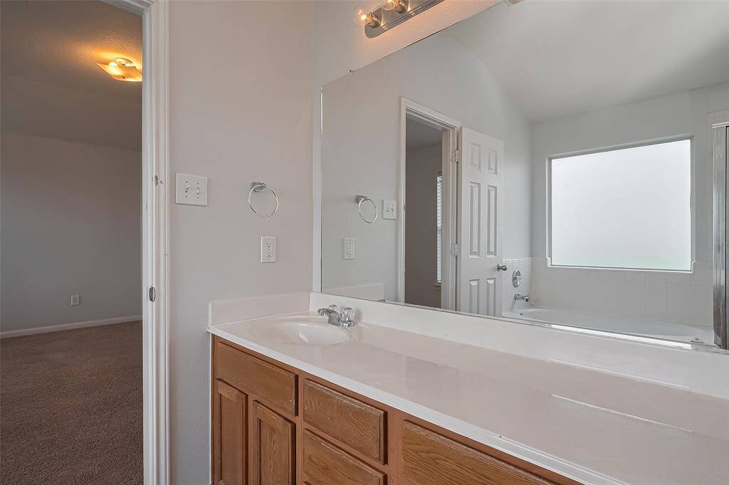 This photo shows a spacious bathroom with a large mirror, a single sink vanity with wooden cabinets, and a bathtub. There's a frosted window providing natural light, and an adjacent door leads to a carpeted room.