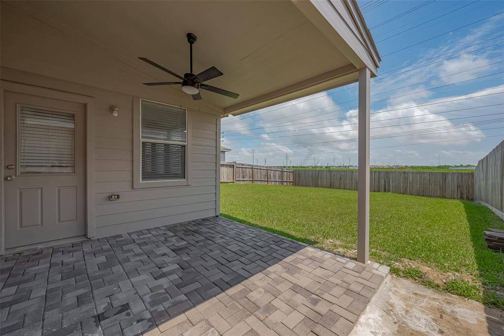 Covered patio with a ceiling fan, featuring a paved surface leading out to a spacious, fenced-in backyard with a well-maintained lawn. Perfect for outdoor relaxation and entertaining.