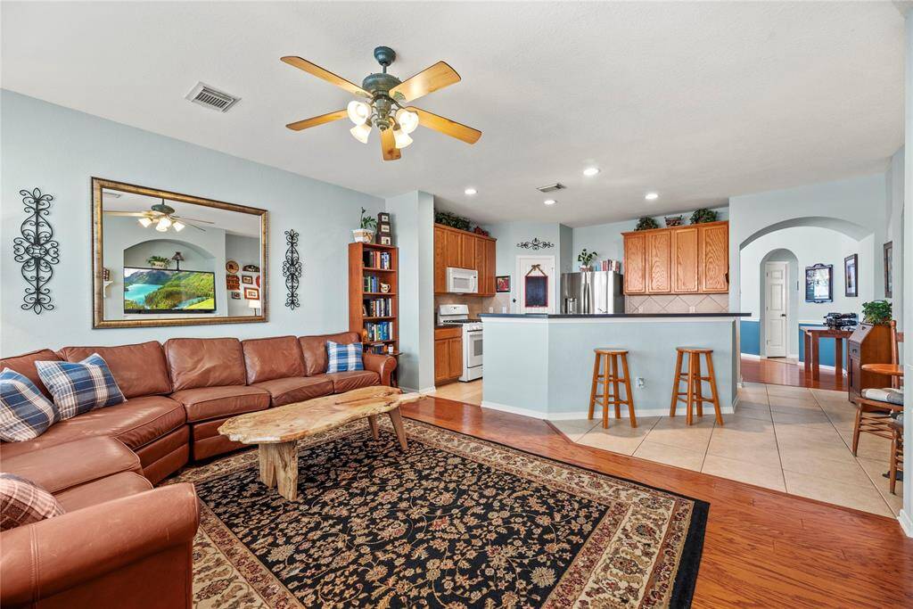 Open-concept living area with an L-shaped leather sofa, a large rug, and a kitchen with wooden cabinets, modern appliances, and a breakfast bar. Soft lighting and ceiling fans add to the relaxing feel.
