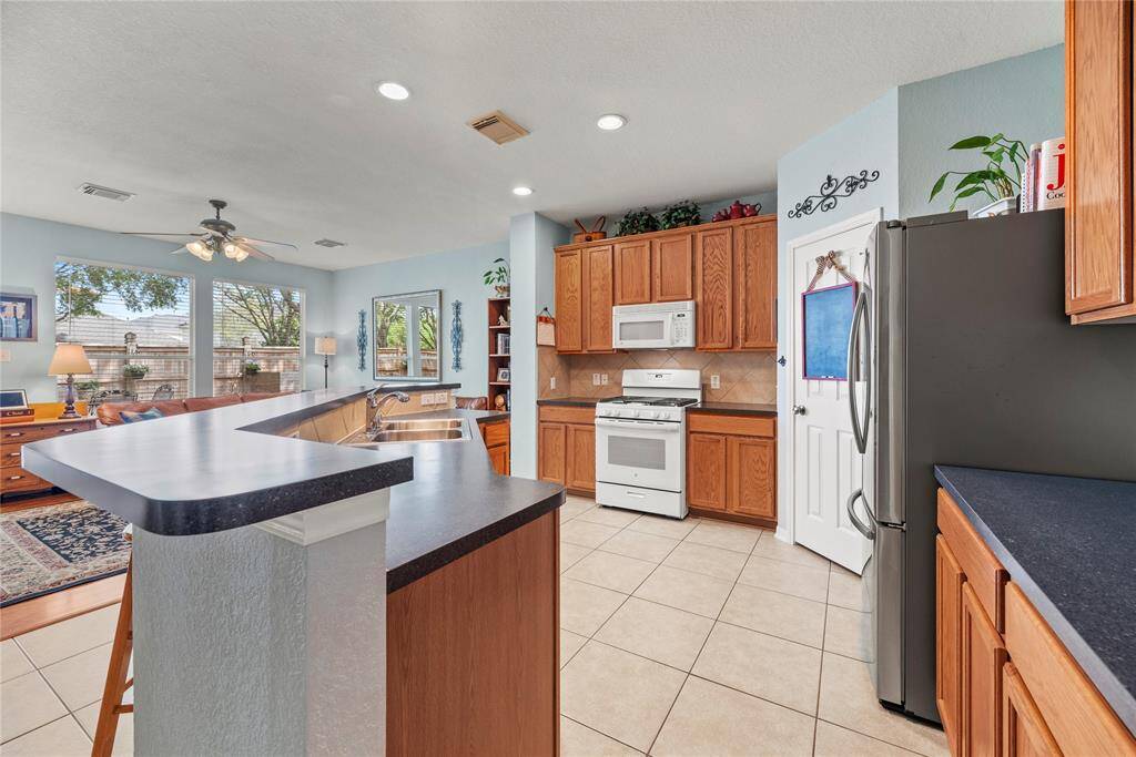 This spacious kitchen features appliances, ample wooden cabinetry, and a large island with a sink. It's open to a bright living area with large windows, creating a warm and inviting space perfect for entertaining.