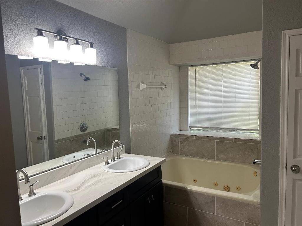 Master bathroom features a double vanity with a marble countertop, modern fixtures, and a large mirror. It includes a spacious bathtub with tile surround and a window with blinds for natural light and view of the back yard.
