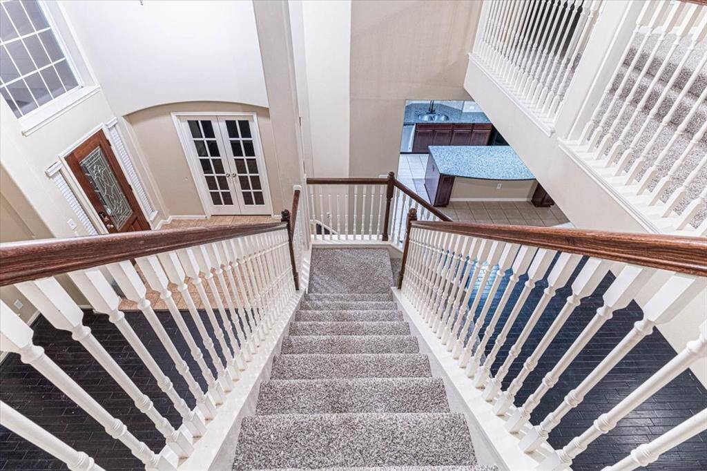 Elegant staircase with a wooden handrail and carpeted steps. It leads down to an open area with a view of a modern kitchen featuring a large island and tiled floor.