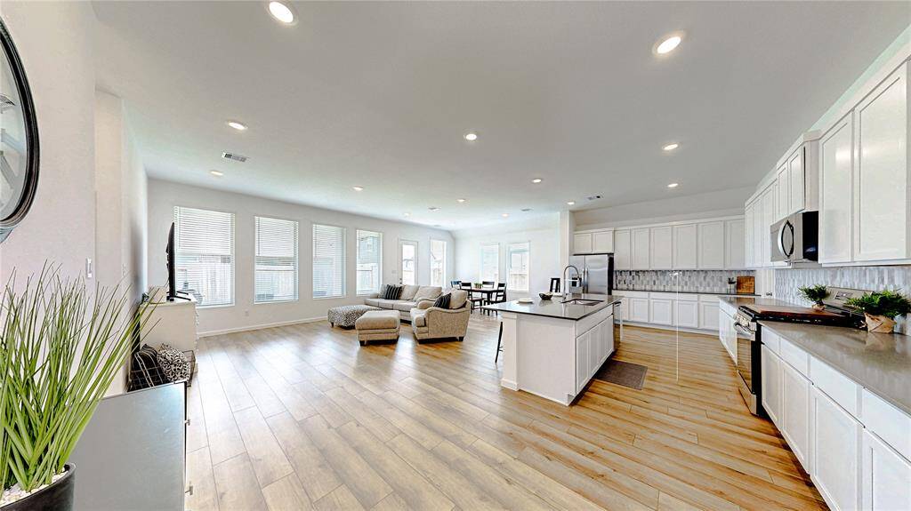 This spacious open-concept living area features a modern kitchen with ample white cabinetry and a large island, seamlessly flowing into a bright living room with abundant natural light from multiple windows. The vinyl plank floors add warmth and elegance, making it perfect for entertaining.