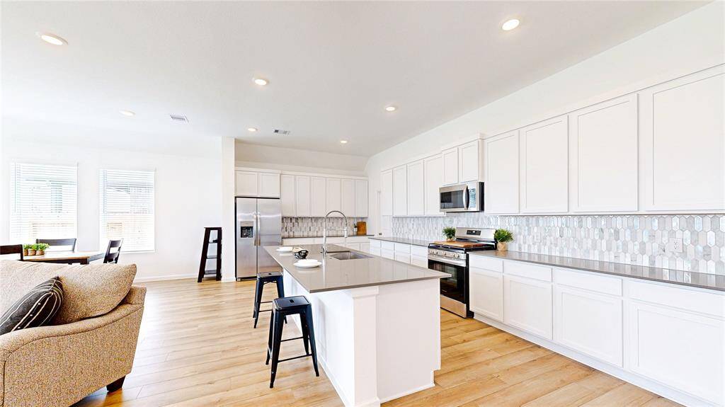 This kitchen features modern white cabinetry, stainless steel appliances, and a stylish tile backsplash. It includes a central island with seating, open to a bright living area with vinyl plank flooring and large windows for natural light.