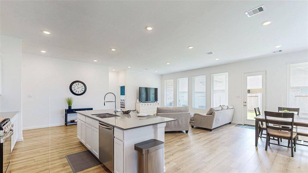 This photo showcases a bright, open-concept living space featuring a modern kitchen with an island, spacious living area, and a dining area. Large windows provide plenty of natural light, and the room has a clean, contemporary feel with light vinyl plank flooring and neutral colors.
