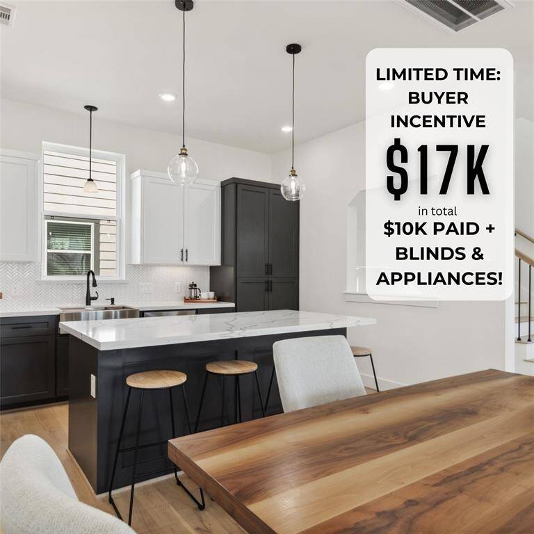 his modern kitchen features sleek black and white cabinetry, a central island with barstools, and pendant lighting. The open layout flows into a dining area with a wooden table, and a staircase leads to the upper level. Large windows provide natural light, creating a bright and inviting space.