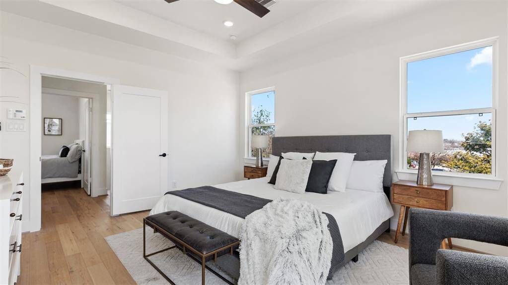 Bright and inviting bedroom featuring a comfortable bed with a gray upholstered headboard, stylish side tables with lamps, and large windows allowing natural light. The room has wooden flooring and a cozy seating area, creating a warm and modern atmosphere.