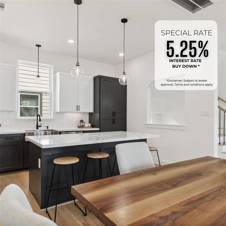 This modern kitchen features sleek black and white cabinetry, a central island with barstools, and pendant lighting. The open layout flows into a dining area with a wooden table, and a staircase leads to the upper level. Large windows provide natural light, creating a bright and inviting space.