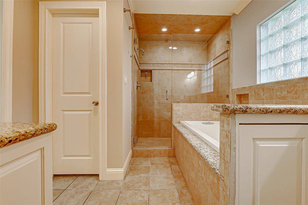 Welcome to your serene primary bathroom, featuring a relaxing soaking tub, a custom walk-in shower, neutral tile floors, and a spacious vanity with elegant granite countertops.