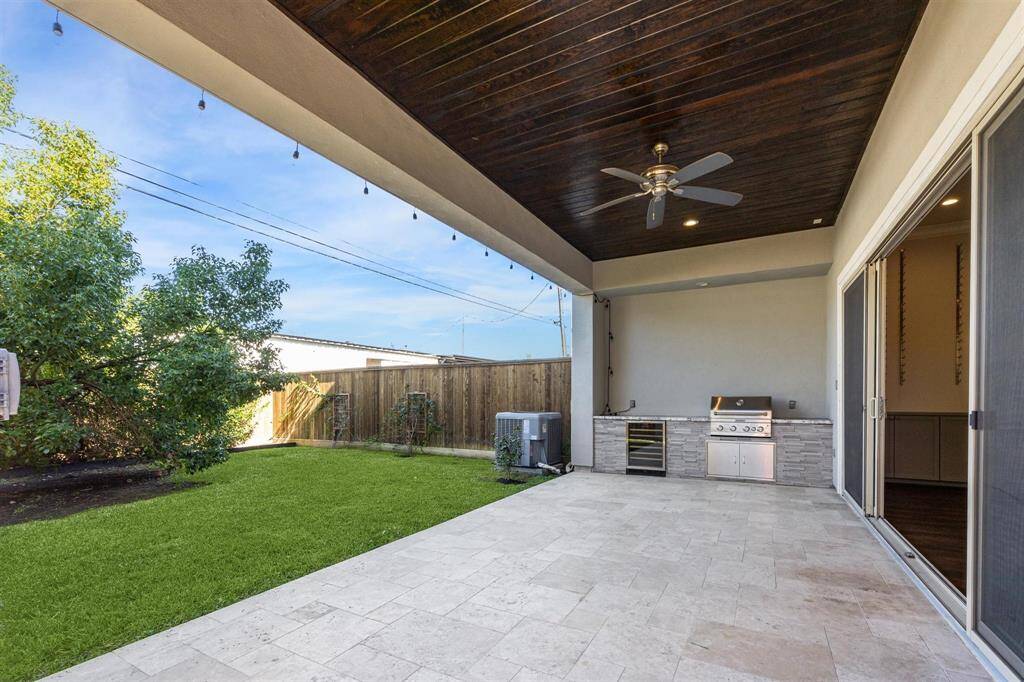 The covered patio offers an outdoor kitchen with a built-in gas grill, beverage fridge, and stone countertops under a dark-stained wood ceiling with recessed lighting and a ceiling fan.