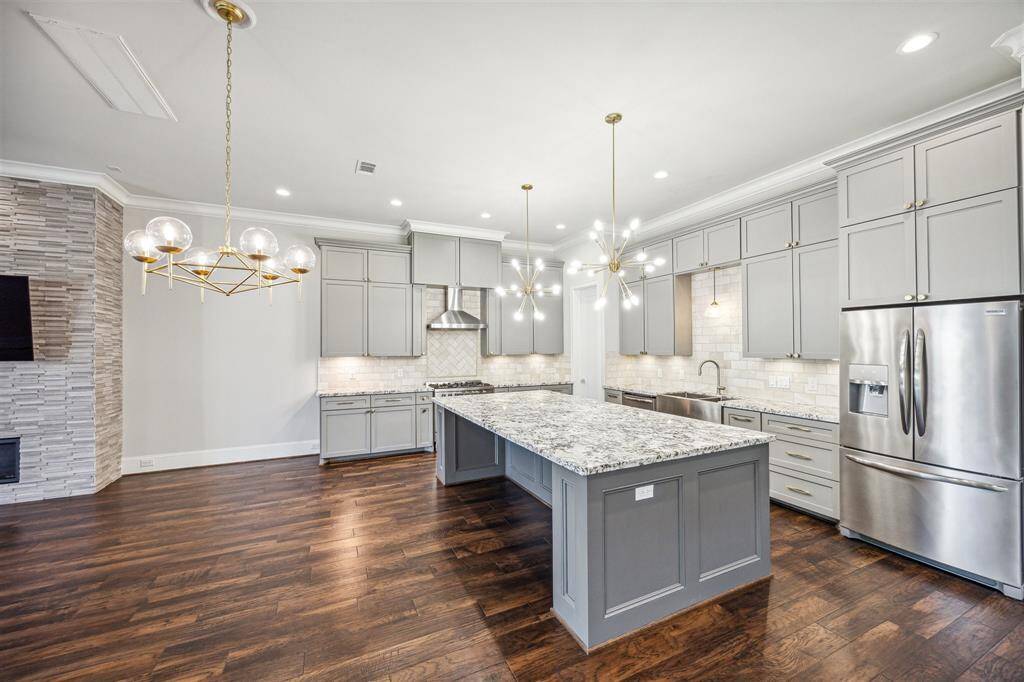 A spacious kitchen with a massive granite island, custom gray cabinetry, brushed gold hardware, and premium stainless steel appliances, seamlessly flowing into the dining area.