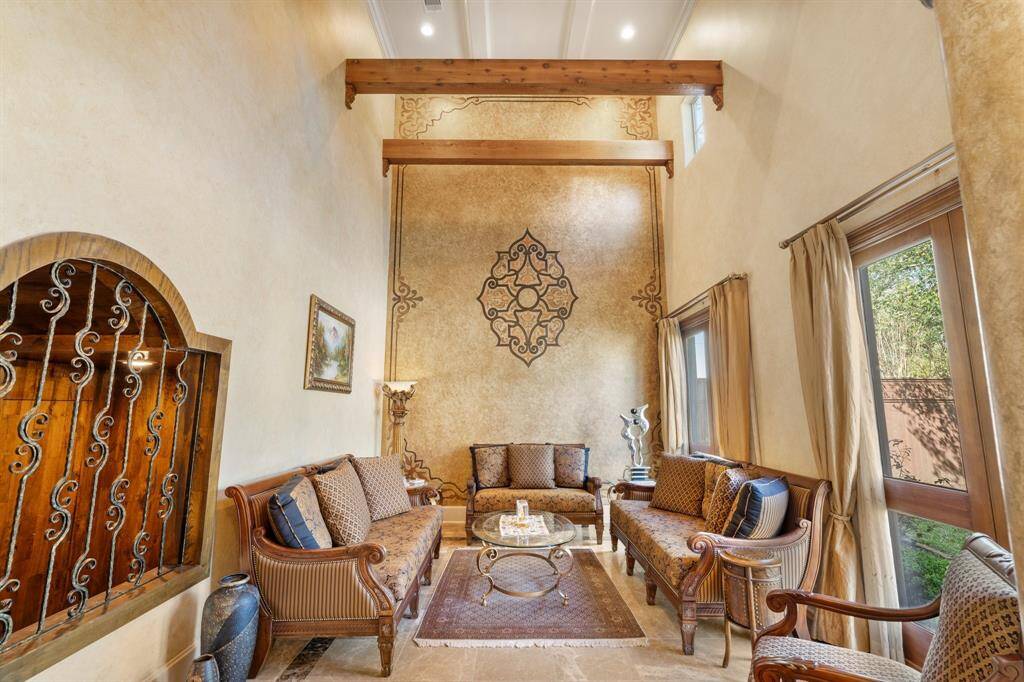 This impressive FORMAL LIVING ROOM boasts a soaring 21-foot double-height ceiling with accent wood beams and a coffered ceiling treatment. The hand-painted textured walls and a stunning hand-painted accent wall add character, while marble floors with intricate inlays elevate the room. Large windows with drapes fill the room with natural light, complemented by recessed can lighting and double crown molding. A decorative iron opening offers a glimpse into the wine cellar for a touch of sophistication.