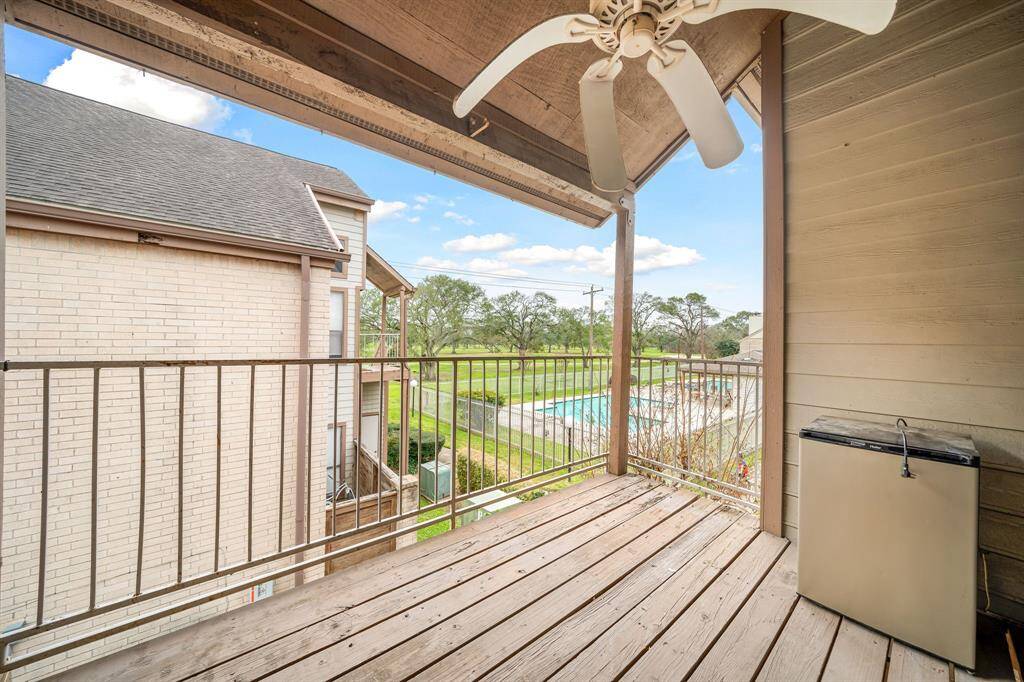 A cozy balcony with wooden flooring, a ceiling fan, and a view of a swimming pool and greenery, perfect for relaxation.