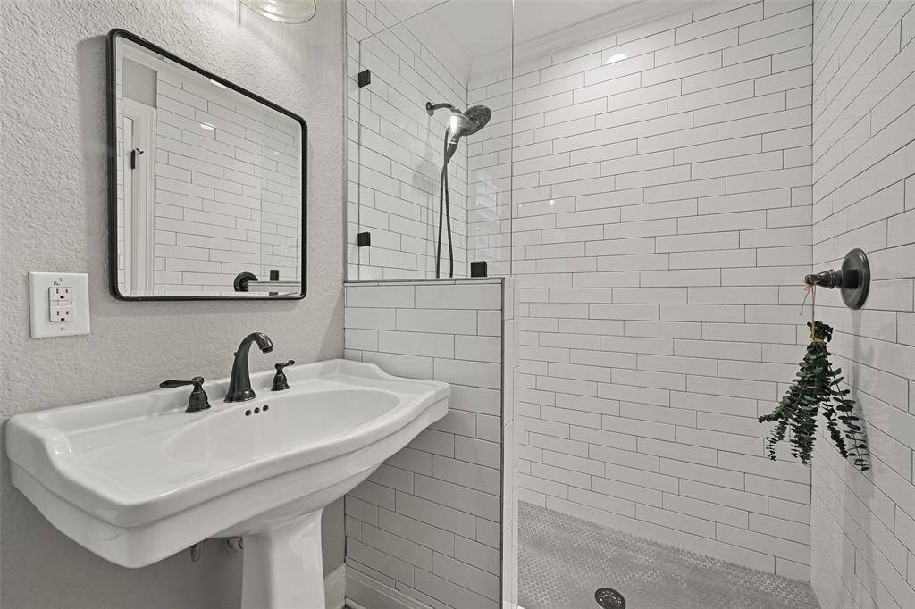 This full bathroom services the gameroom as well as backyard pool guests, featuring tile floors, a pedestal sink topped by a framed mirror, stylishly updated lighting, and a walk-in shower with a handheld showerhead and a subway tile enclosure.