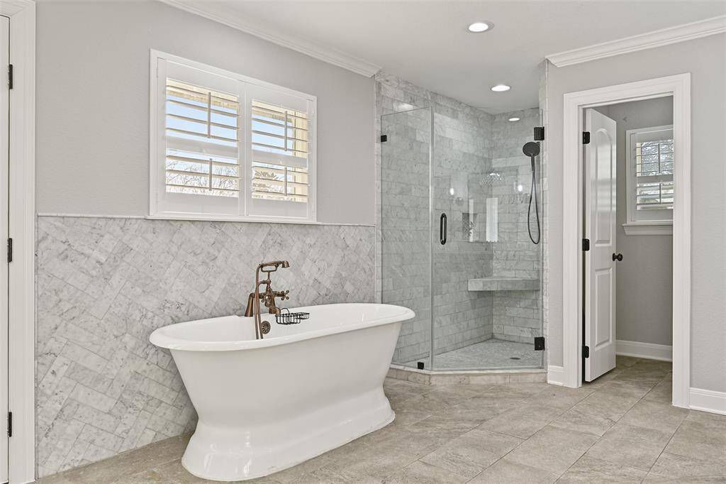 Relaxation awaits beneath the deep waters of this freestanding soaker tub with a wall-mounted bronze faucet & handheld sprayer and a marble tile splash wall that extends into the seamless glass shower with a bench seat.