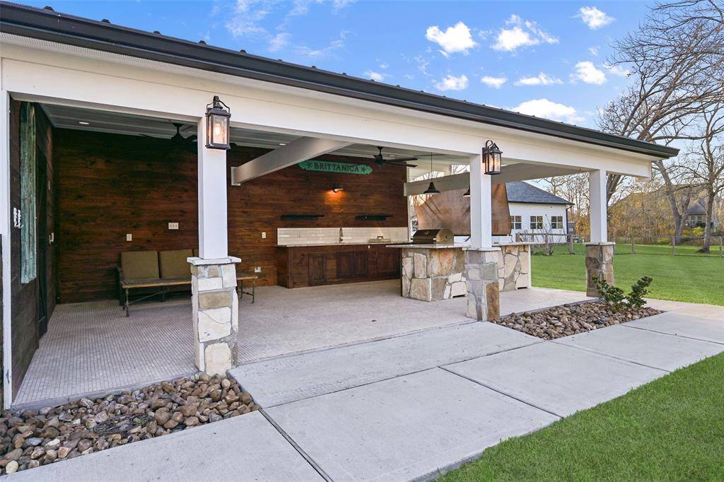 A closer look at the bonus patio highlights the outdoor kitchen with a stone and wood base, granite countertops, a subway tile backsplash, modern lighting along with recessed lighting, a wood slat accent wall, a single basin sink, and a gas grill. To the left, there is a private sauna!