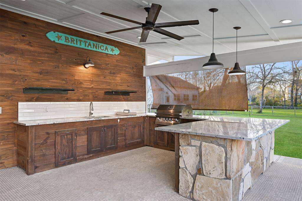 A closer look at the bonus patio highlights the outdoor kitchen with a stone and wood base, granite countertops, a subway tile backsplash, modern lighting along with recessed lighting, a wood slat accent wall, a single basin sink, and a gas grill. To the left, there is a private sauna!