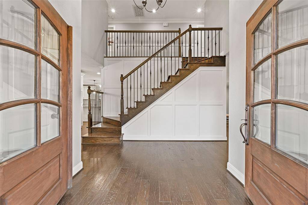 A pair of farmhouse-style front doors open from the covered front porch into this warm and inviting foyer that features designer lighting, neutral paint, engineered wood floors, and a handsome staircase with wood treads and handrails and scrolled iron balusters.