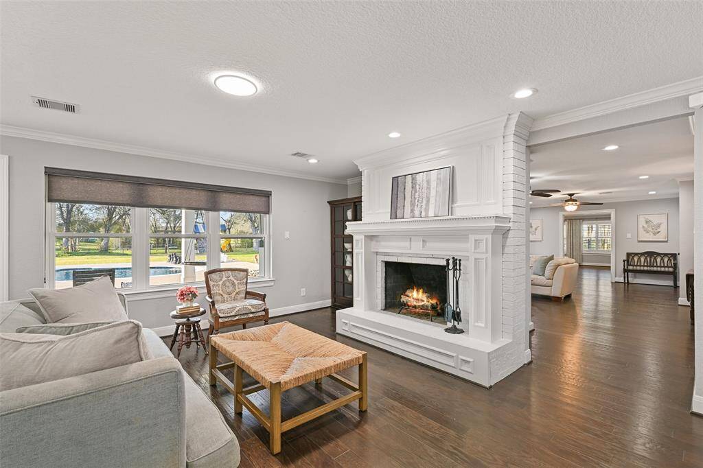 Continue into the heart of the home where the living space is broken up into multiple gathering areas. In this space, note the painted brick fireplace with a milled wood mantle, recessed lighting, crown molding, wood floors, and trio of windows with pull-down shades and pool views.