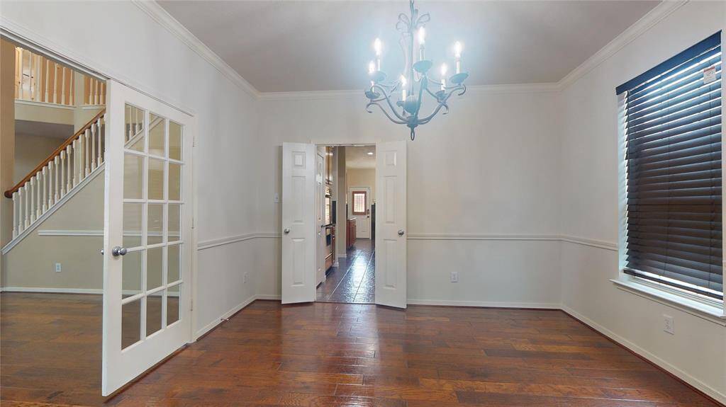 This room features hardwood flooring, a stylish chandelier, and ample natural light from a large window with blinds. French doors lead to a staircase, and there's an entryway to a kitchen area.
